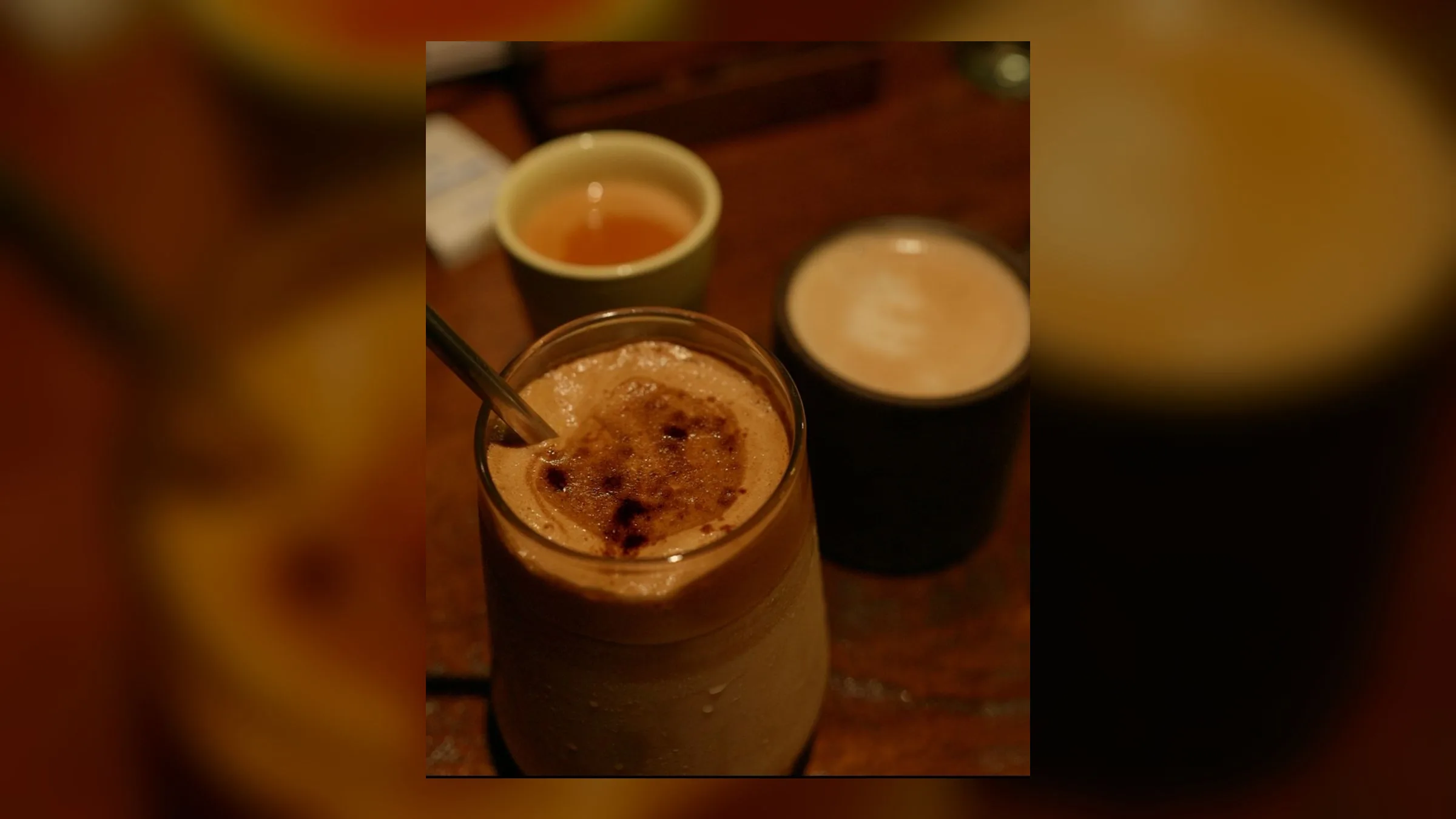 A tall glass of frothy cold coffee dusted with cocoa, a metal straw resting in it, cups of tea and coffee on the warm wooden table behind
