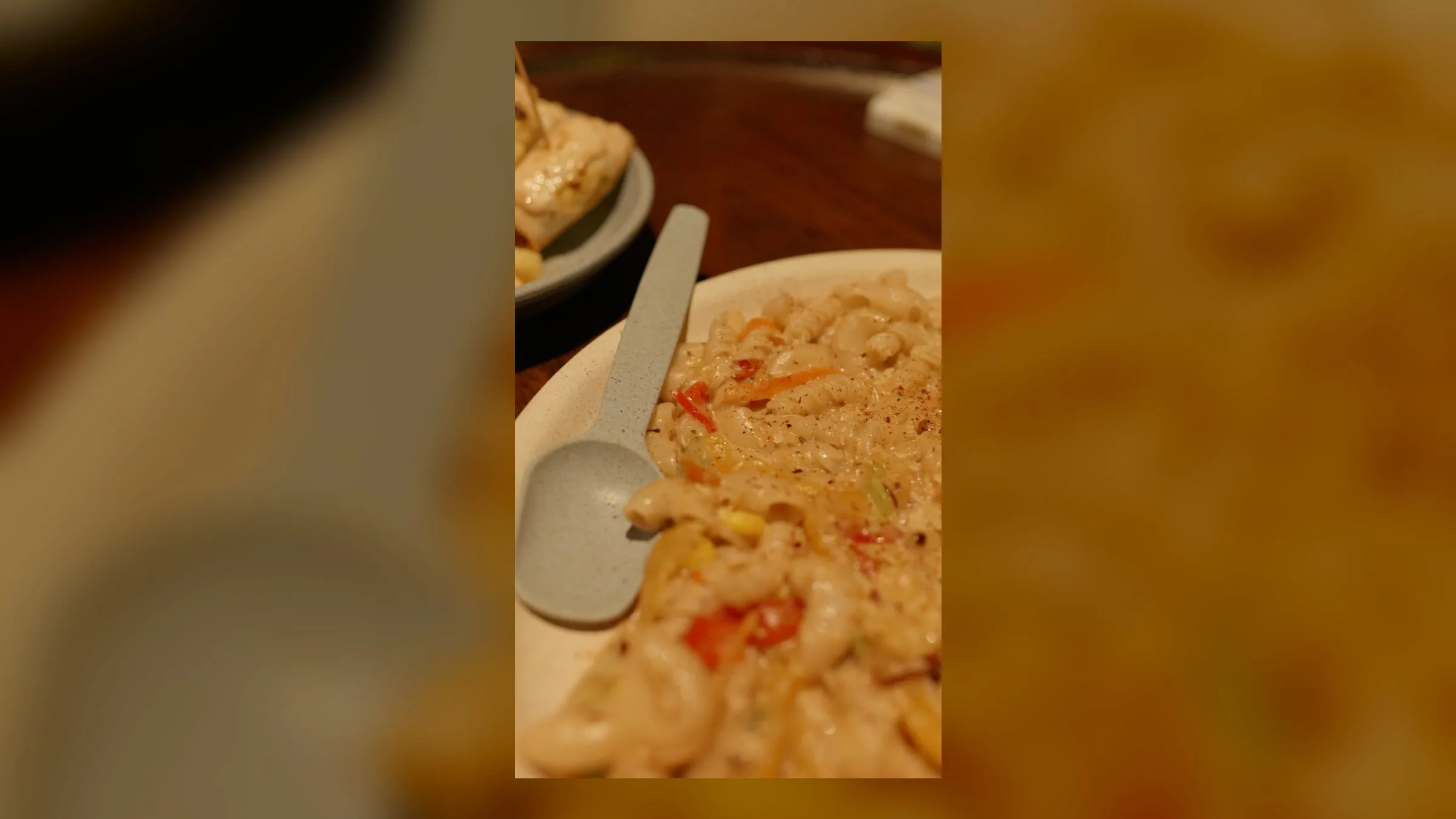 A creamy plate of pasta tossed with colourful peppers and sweetcorn, a ceramic spoon on the rim, skewers on a plate behind